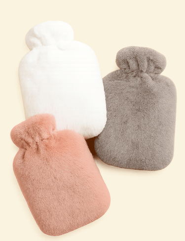 Three Warmweave™ hot water bottles in soft pink, grey, and white covers, displayed on a warm neutral background.